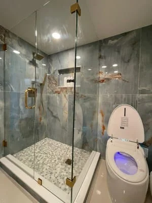 Frameless glass shower with marble tile walls, pebble floor, and brass fixtures in renovated bathroom